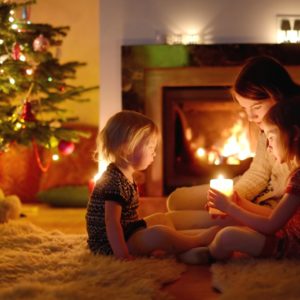 Young mother and her two little daughters sitting by a fireplace holding a candle in a cozy dark living room on Christmas eve